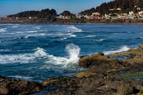 Waves crash against rocky shore with a coastal town in the background under a clear blue sky.