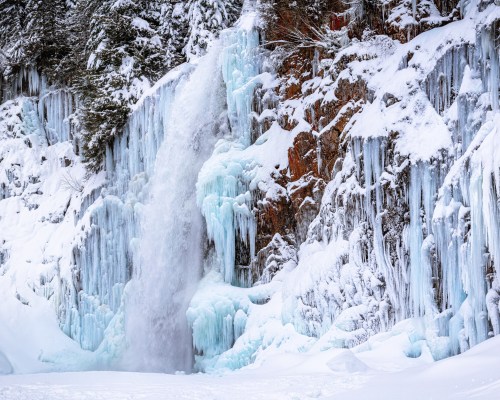 A frozen waterfall cascading down icy cliffs, surrounded by snow-covered trees and a winter landscape.