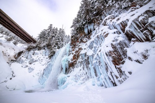 A frozen waterfall cascading down a snowy cliff, with icicles hanging and a bridge above in a winter landscape.