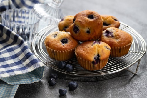 A wire rack holds freshly baked blueberry muffins, with a few blueberries scattered nearby and a checkered cloth in the background.