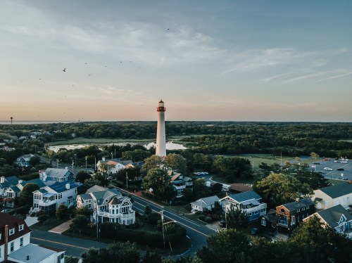 Aerial view of a coastal town with a lighthouse, surrounded by trees and houses, under a clear sky at sunset.