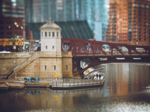 A small dock on a river with a bridge in the background, surrounded by city buildings and reflections in the water.