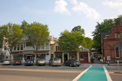 A charming small-town street with shops, trees, and parked cars under a clear blue sky.