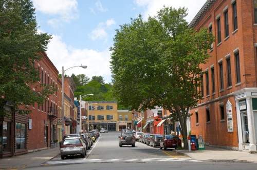 A quaint street lined with brick buildings and trees, featuring parked cars and shops under a partly cloudy sky.