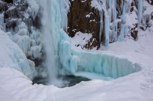 A frozen waterfall cascading into a snowy, icy pool surrounded by frosted rocks and ice formations.