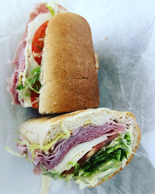 A close-up of two halves of a sandwich with layers of ham, turkey, lettuce, and tomato on a soft bread roll.