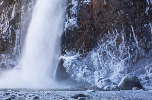 A frozen waterfall cascading down a rocky cliff, surrounded by icy formations and a rocky riverbed.