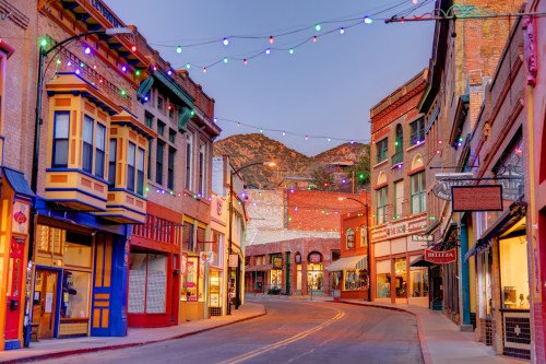 Colorful buildings line a quiet street adorned with festive lights, set against a mountain backdrop at dusk.