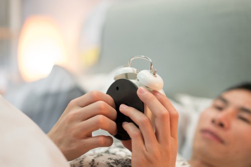 A person in bed adjusts an alarm clock, with soft lighting in the background.