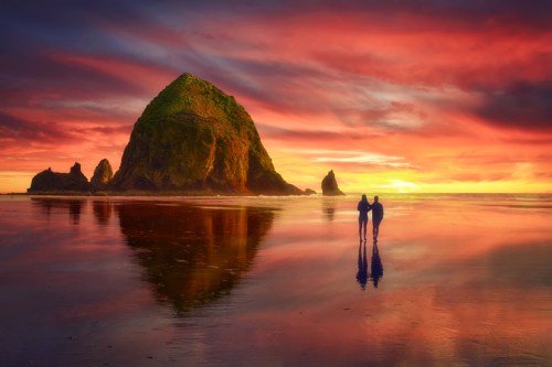 A couple stands on a beach at sunset, with vibrant colors reflecting on the water and a large rock formation in the background.