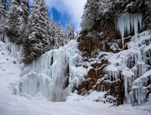 A frozen waterfall surrounded by snow-covered trees and rocky cliffs under a blue sky.