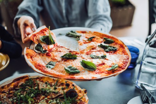 A person holds a pizza topped with fresh basil, ready to serve, with another pizza visible on the table.