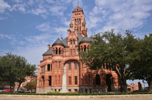 Historic red brick building with ornate architecture, surrounded by trees and a statue in front, under a blue sky.