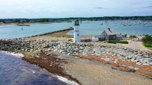 A scenic view of a lighthouse by the shore, surrounded by boats and rocky coastline under a clear sky.