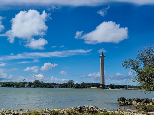 A tall tower stands by a calm lake under a blue sky with fluffy clouds, surrounded by greenery and distant buildings.