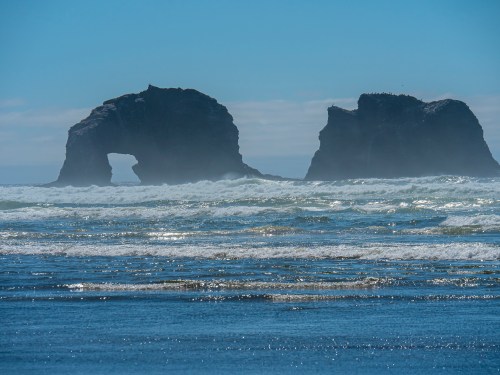 Two large rock formations rise from the ocean, partially shrouded in mist, with waves gently lapping at the shore.