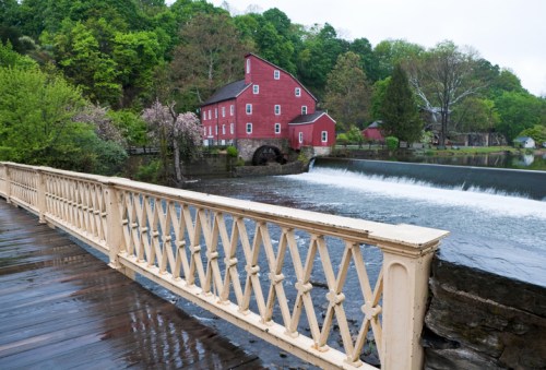 A red historic mill beside a river, with a wooden railing in the foreground and lush greenery in the background.