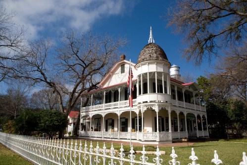 A large, historic white house with a dome, surrounded by trees and a white picket fence under a blue sky.