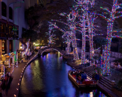 A serene night scene of a river lined with colorful holiday lights and trees, with a boat gliding through the water.