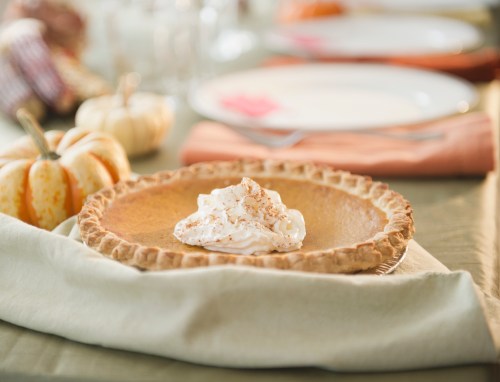 A pumpkin pie topped with whipped cream, set on a table with small pumpkins and empty plates in the background.