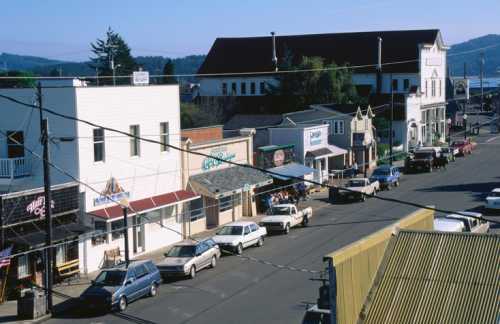 A small town street lined with shops and parked cars, surrounded by hills and a clear blue sky.