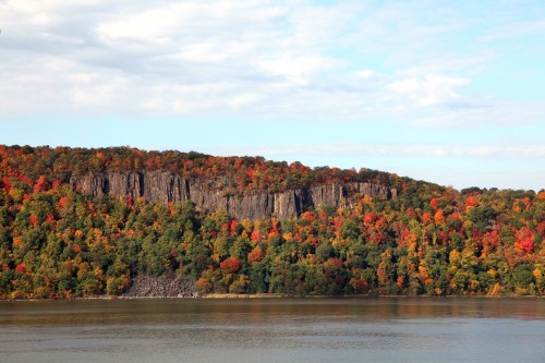 Vibrant autumn foliage on a rocky hillside overlooking a calm river under a partly cloudy sky.