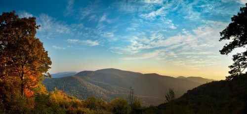 A panoramic view of rolling mountains under a blue sky, with autumn foliage in the foreground.