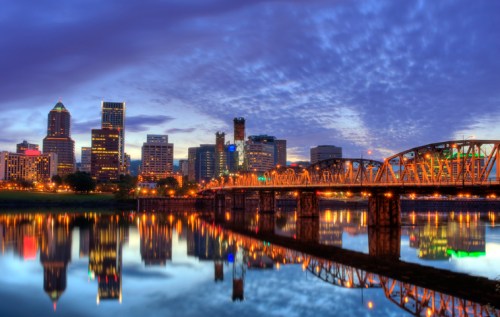 City skyline at dusk with illuminated buildings and a bridge reflected in the water below.