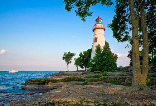 A white lighthouse with a red top stands by the shore, surrounded by trees and rocky terrain, with a boat in the distance.