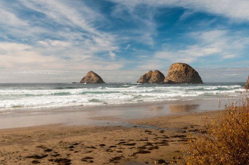 A serene beach scene with three rocky formations in the ocean under a cloudy sky, with gentle waves lapping at the shore.