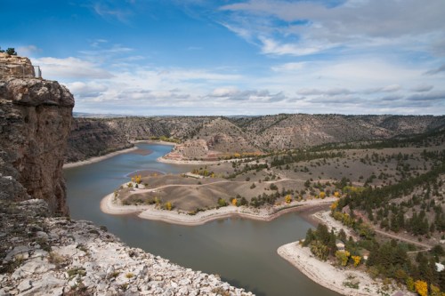 A scenic view of a winding river surrounded by hills and trees under a partly cloudy sky.