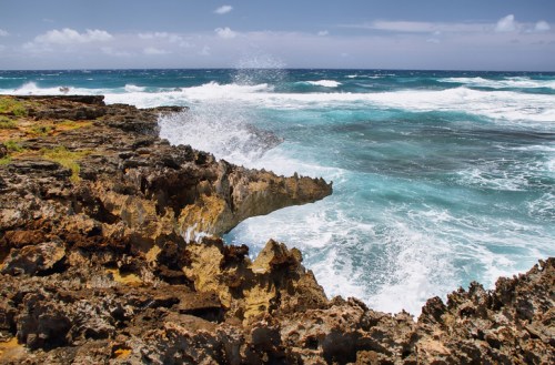 Waves crash against rocky coastline under a bright blue sky, creating a scenic ocean view.