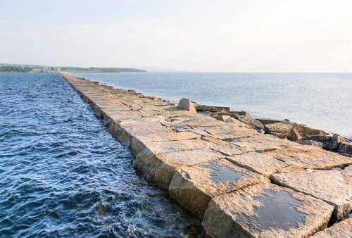 A stone jetty extends into calm waters, leading towards a distant shoreline under a clear sky.
