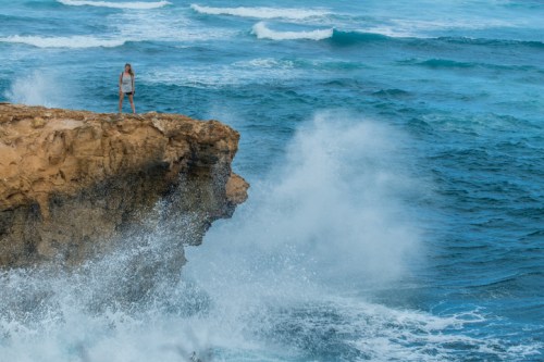 A person stands on a rocky cliff overlooking crashing waves in a vibrant blue ocean.