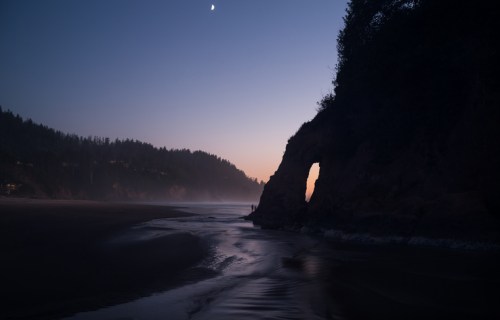 A serene beach at dusk, featuring a rocky arch, gentle waves, and a crescent moon in the sky.