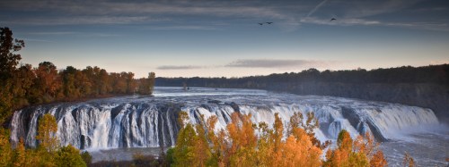 A panoramic view of a waterfall surrounded by trees, with birds flying in the sky above.