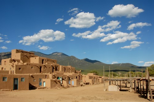 Adobe buildings with blue doors under a bright sky, surrounded by mountains and scattered clouds. Desert landscape.