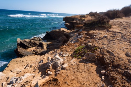 Rocky coastline with waves crashing against cliffs under a clear blue sky. Sparse vegetation along the shore.