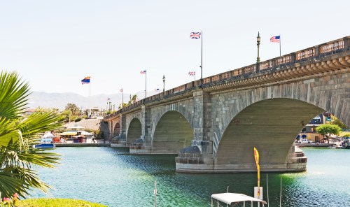 A stone bridge with flags over a calm waterway, surrounded by palm trees and buildings in the background.