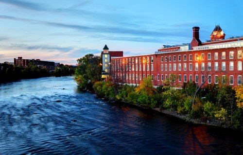 A brick building along a river at sunset, with colorful trees and a clear blue sky reflecting on the water.