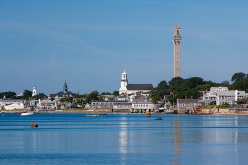 A scenic view of a coastal town with a tall tower, church steeples, and calm waters under a clear blue sky.