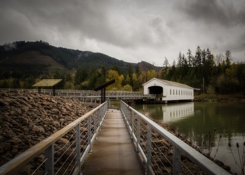 A wooden walkway leads to a white covered bridge over a calm river, surrounded by mountains and trees under a cloudy sky.