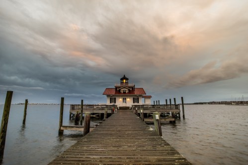 A wooden pier leads to a lighthouse on calm waters, under a dramatic sky with clouds at sunset.