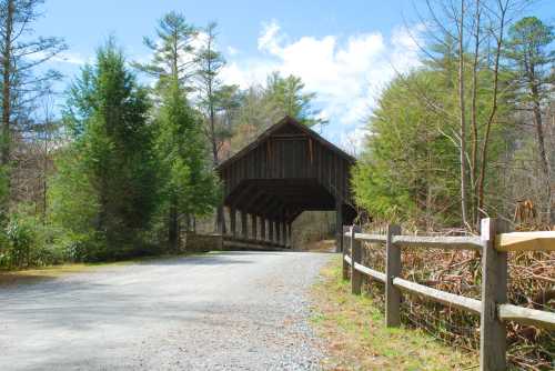 A rustic covered bridge stands over a gravel road, surrounded by trees and a wooden fence under a blue sky.