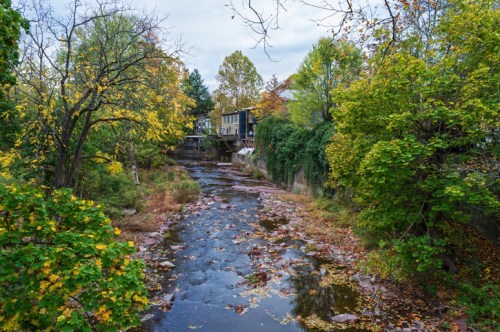 A serene river flows through a lush, green landscape with autumn foliage and a house in the background.