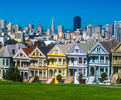 Colorful Victorian houses in front of a city skyline, featuring tall buildings and a clear blue sky.