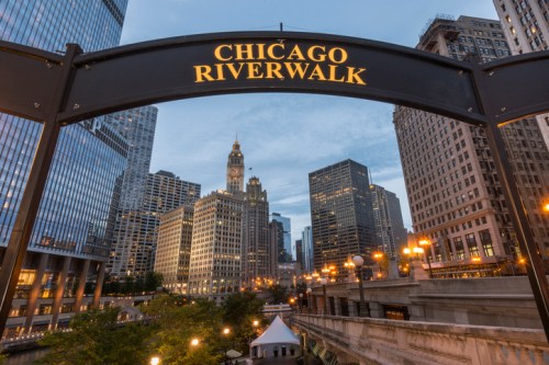 View of the Chicago Riverwalk sign with city skyline and buildings illuminated at dusk.