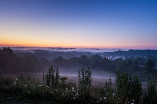A serene landscape at dawn, featuring misty hills and a colorful sky transitioning from night to day.