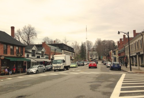 A quaint town street lined with shops, parked cars, and a cloudy sky, creating a charming small-town atmosphere.