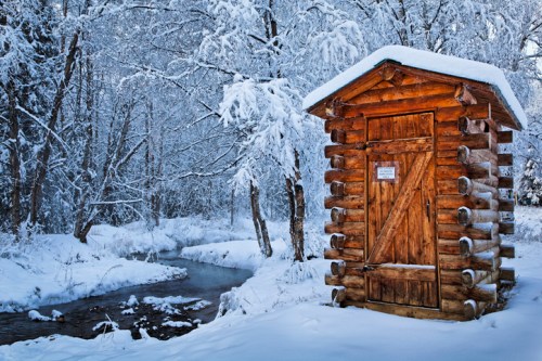 A wooden shed covered in snow, surrounded by frosty trees and a small stream in a winter landscape.